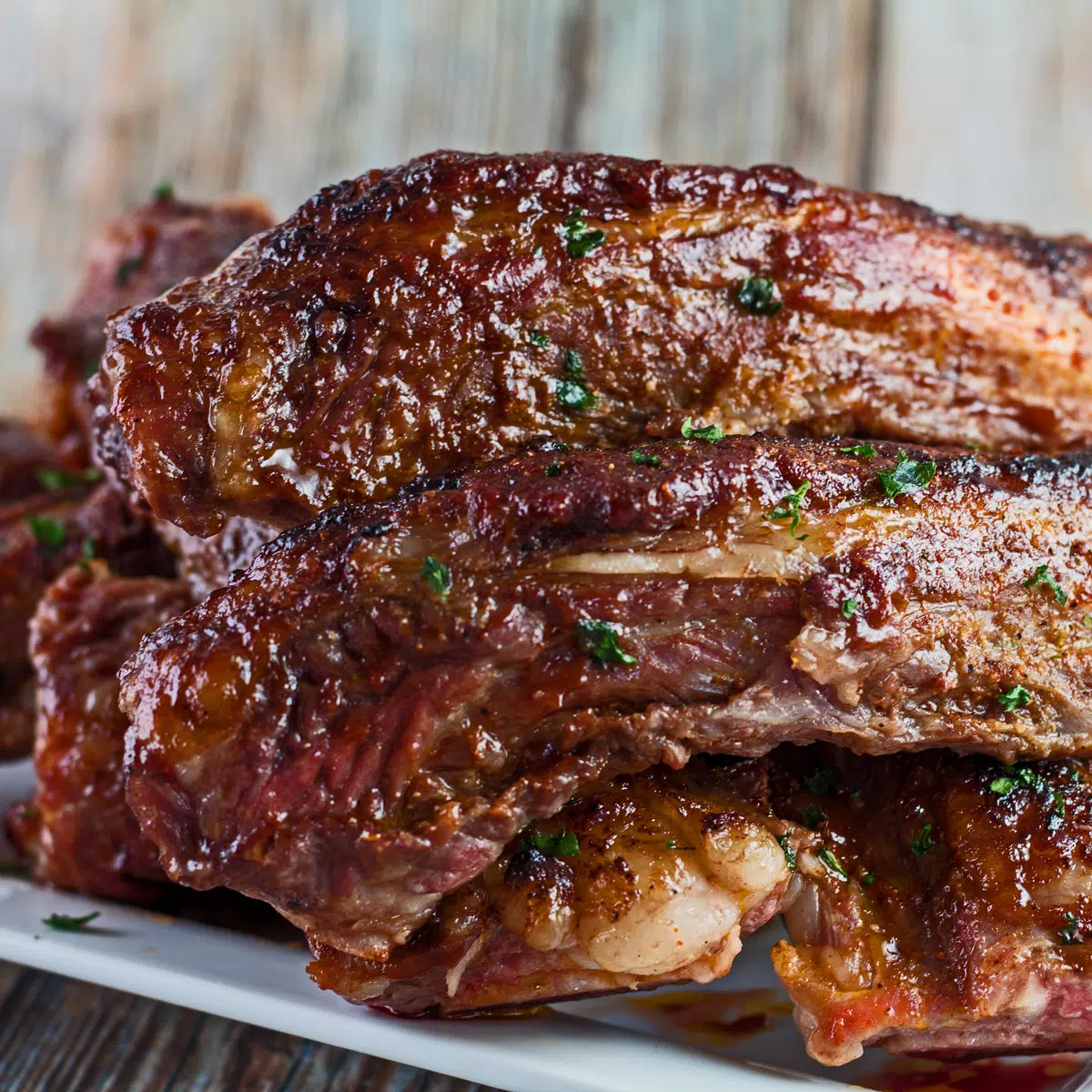 Iga punggung sapi panggang BBQ ditumpuk di piring putih dengan latar belakang biji-bijian kayu.