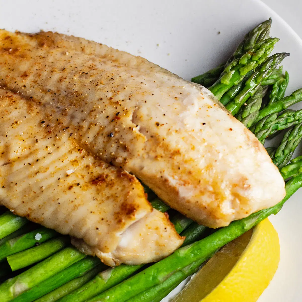 Air fryer tilapia served over asparagis with lemon wedge on white plate.