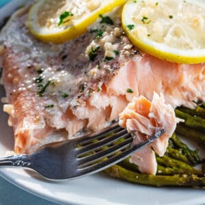 Salmon panggang dalam foil disajikan dengan asparagus di piring putih.