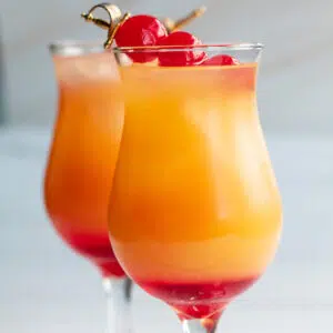A set of two malibu sunrise cocktails in small hurricane glasses with maraschino cherry garnish.