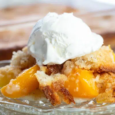 Bisquick peach cobbler served on glass plate with baking dish in background.