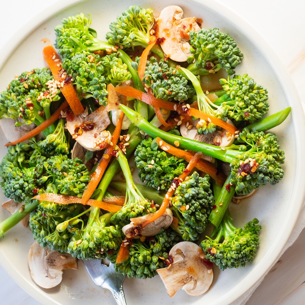 Broccolinisalade op wit bord met champignons, geschaafde wortelen en lichte Aziatische dressing.