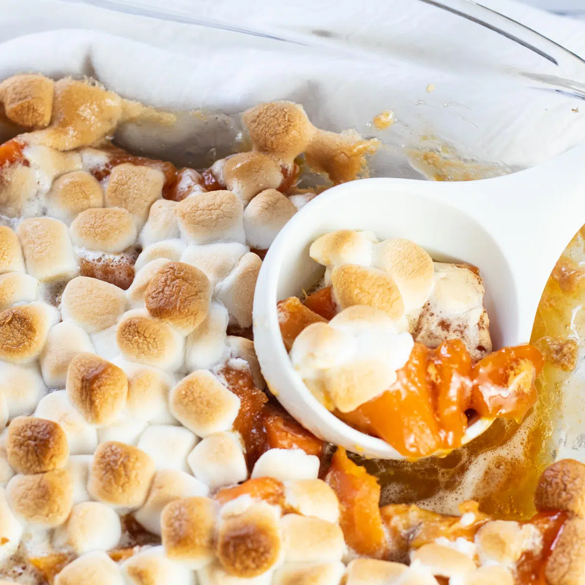Candied yams with marshmallows in glass baking dish with white serving spoon.