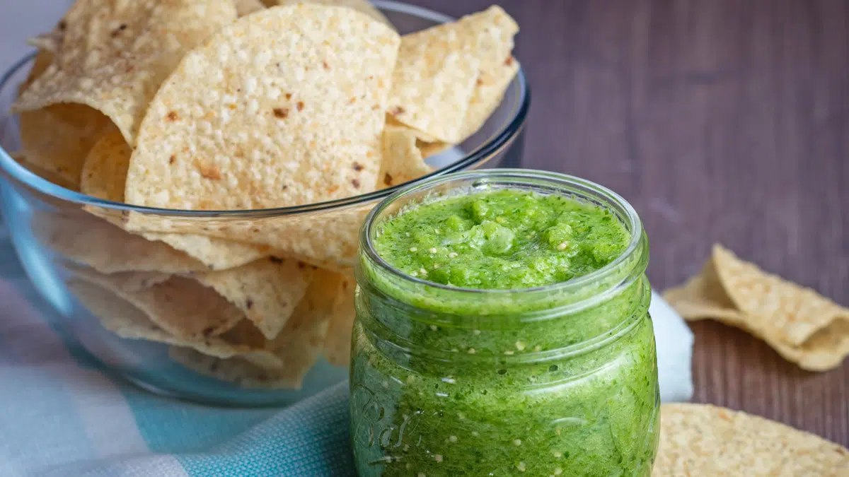 Wide image of salsa verde in a jar with tortilla chips.