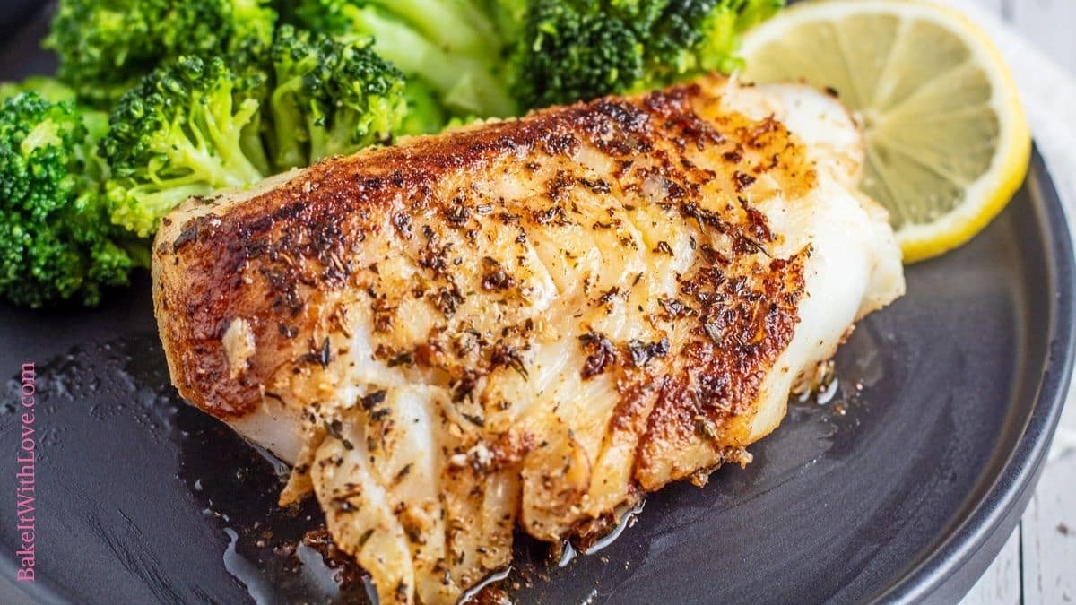 Wide image of blackened cod fish on a black plate with broccoli and a lemon slice.