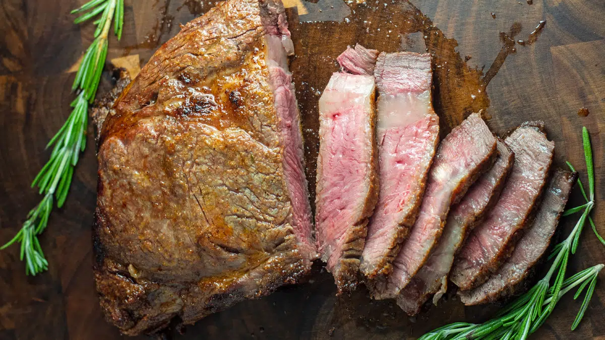 Wide image of a broiled ribeye steak on a wood cutting board sliced.
