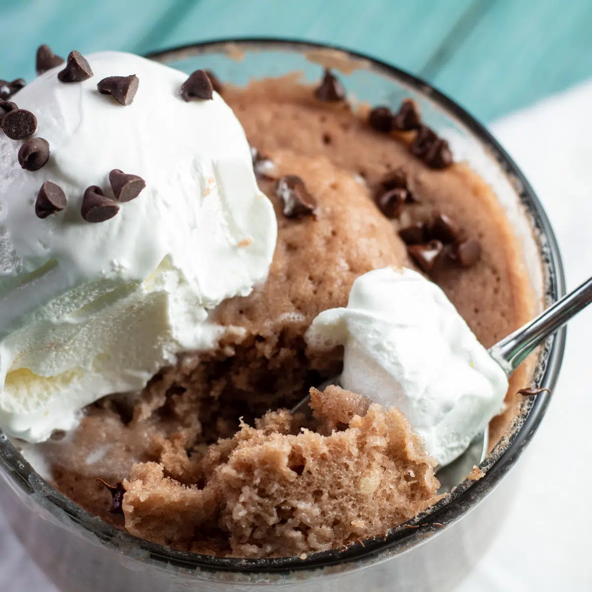 Mug cake in polvere proteica al cioccolato in tazza trasparente con panna montata e mini gocce di cioccolato.