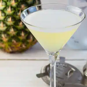 Square close up image of pineapple martini with fresh pineapple behind.