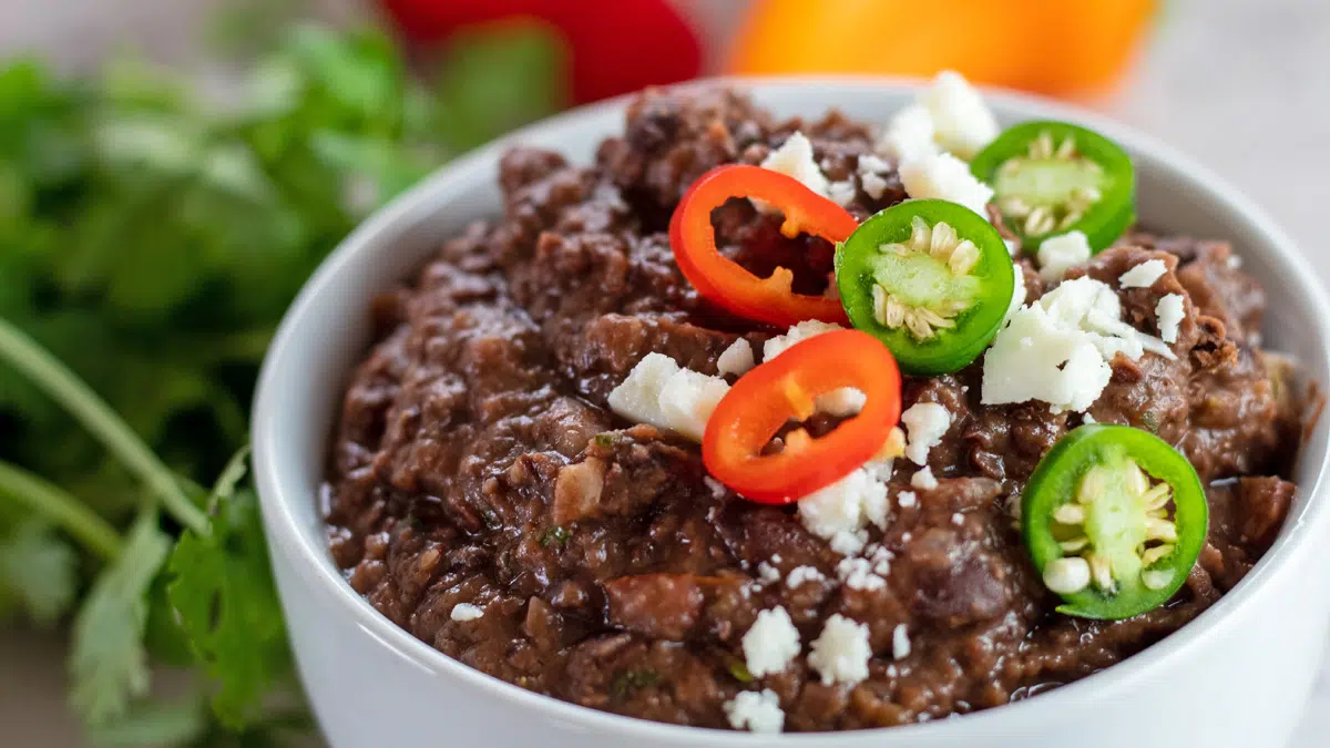 White bowl of refried black beans with garnishes on top.