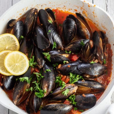 Square image of Spanish mussels in a white sauce pan with lemon slices.