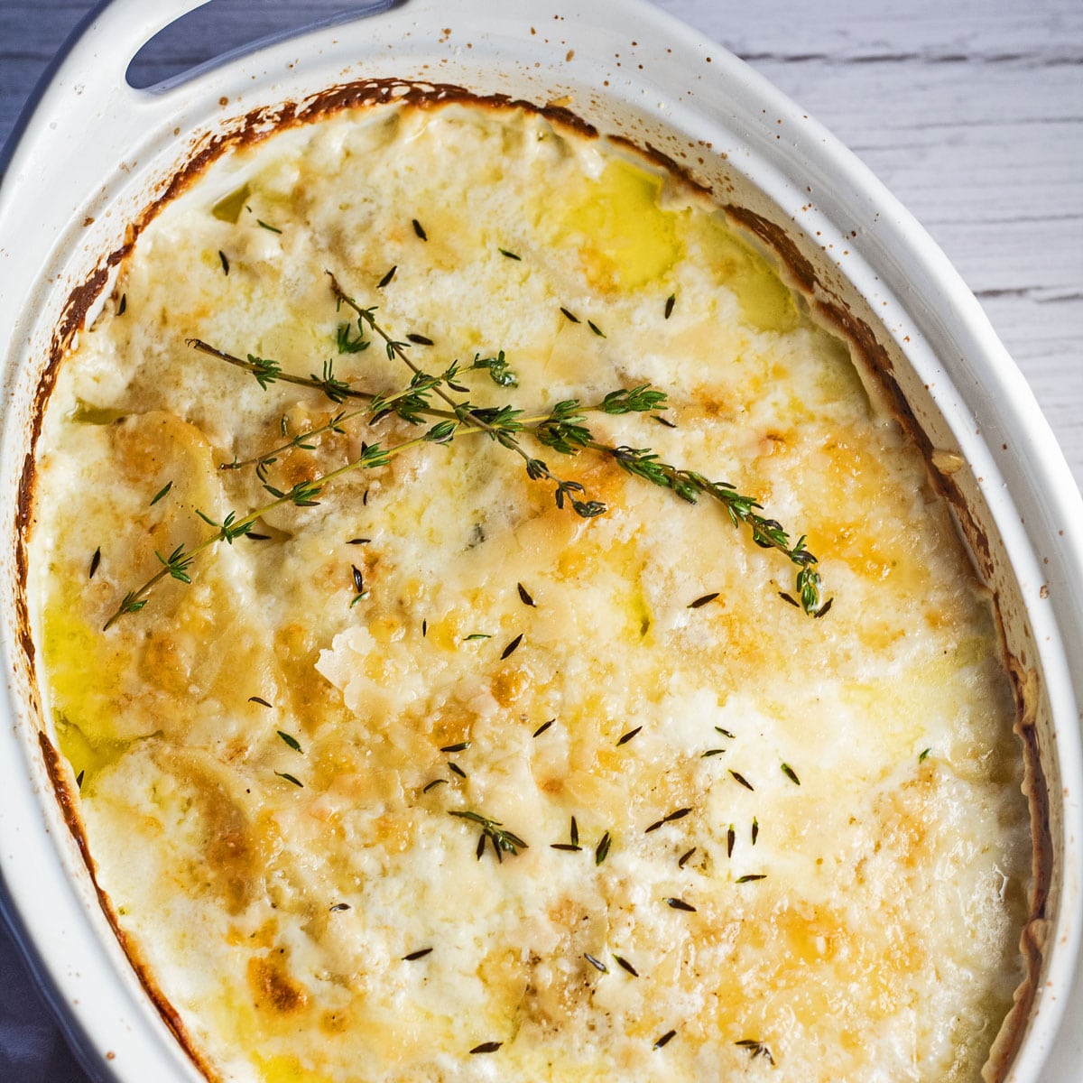 Baked dauphinoise potatoes in white oval casserole dish with fresh thyme garnish.