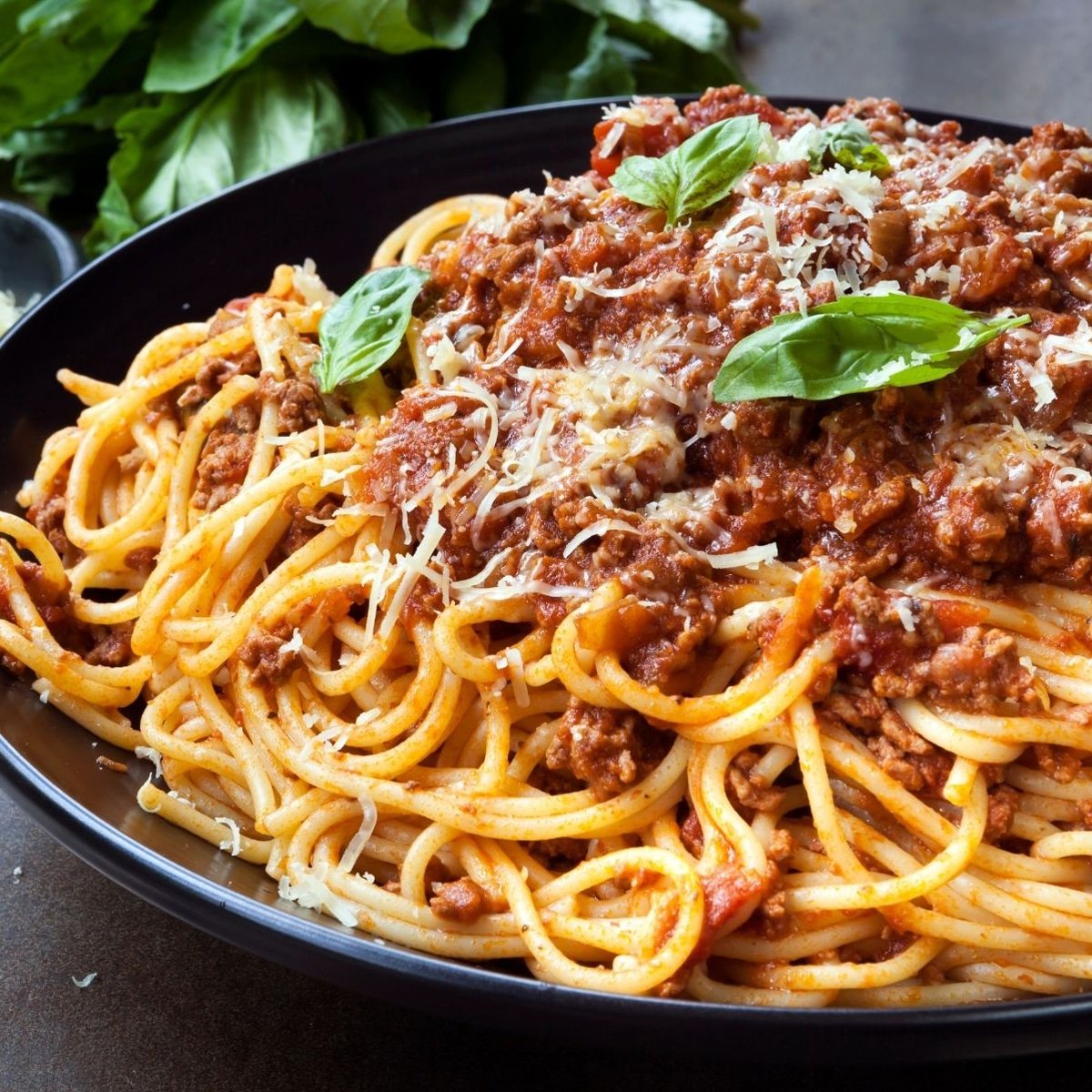 Espaguetis a la boloñesa en un plato negro con parmesano y guarnición de albahaca.