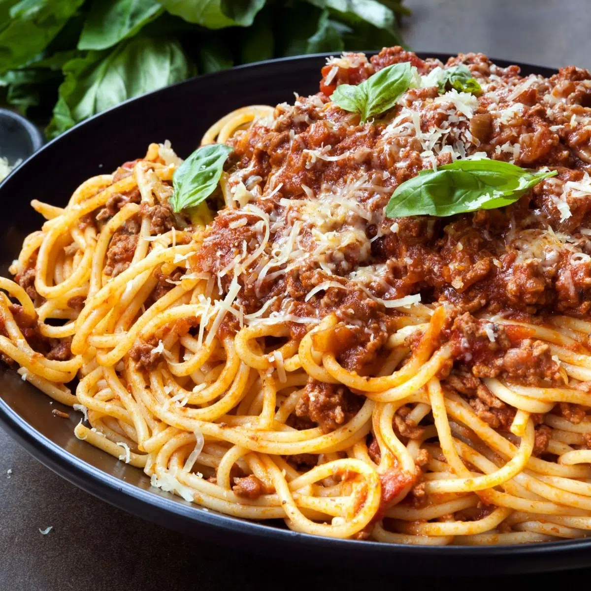 Spaghetti bolognese on black plate with Parmesan and basil garnish.