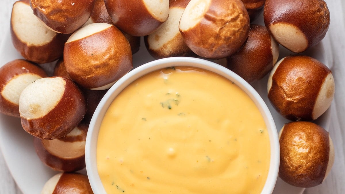 Wide image of cheese dip in a white bowl with pretzels around bowl.