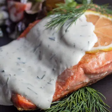 Wide image of salmon with dillsauce on a black plate with lemon slice.