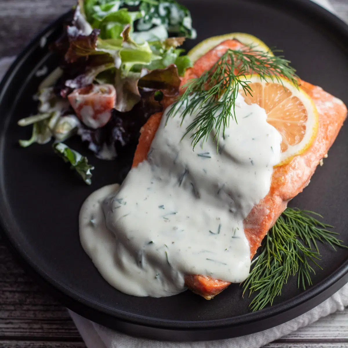 Square image of salmon with dillsauce on a black plate with lemon slice.