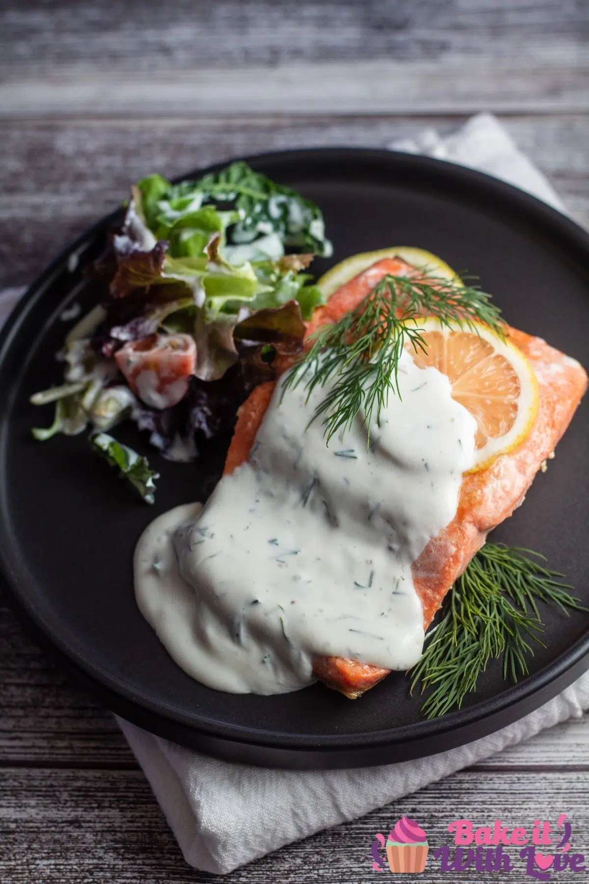 Tall image of salmon with dillsauce on a black plate with lemon slice.