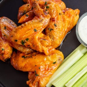 Square image of chicken wings on a black plate with celery.