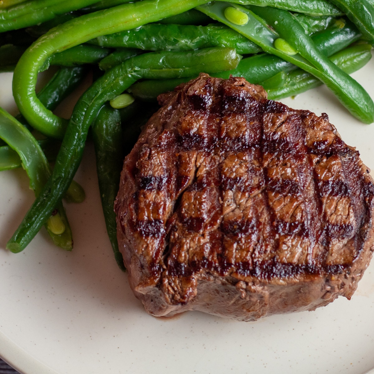 Square image of grilled filet mignon on a plate with green beans.