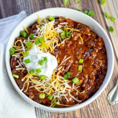 Overhead square image of the ground venison chili served in white bowl with shredded cheese sour cream and green onions.