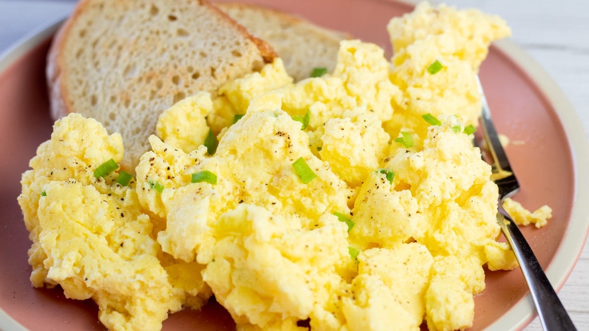 Wide image of scrambled eggs on rose colored plate with toast on the side.
