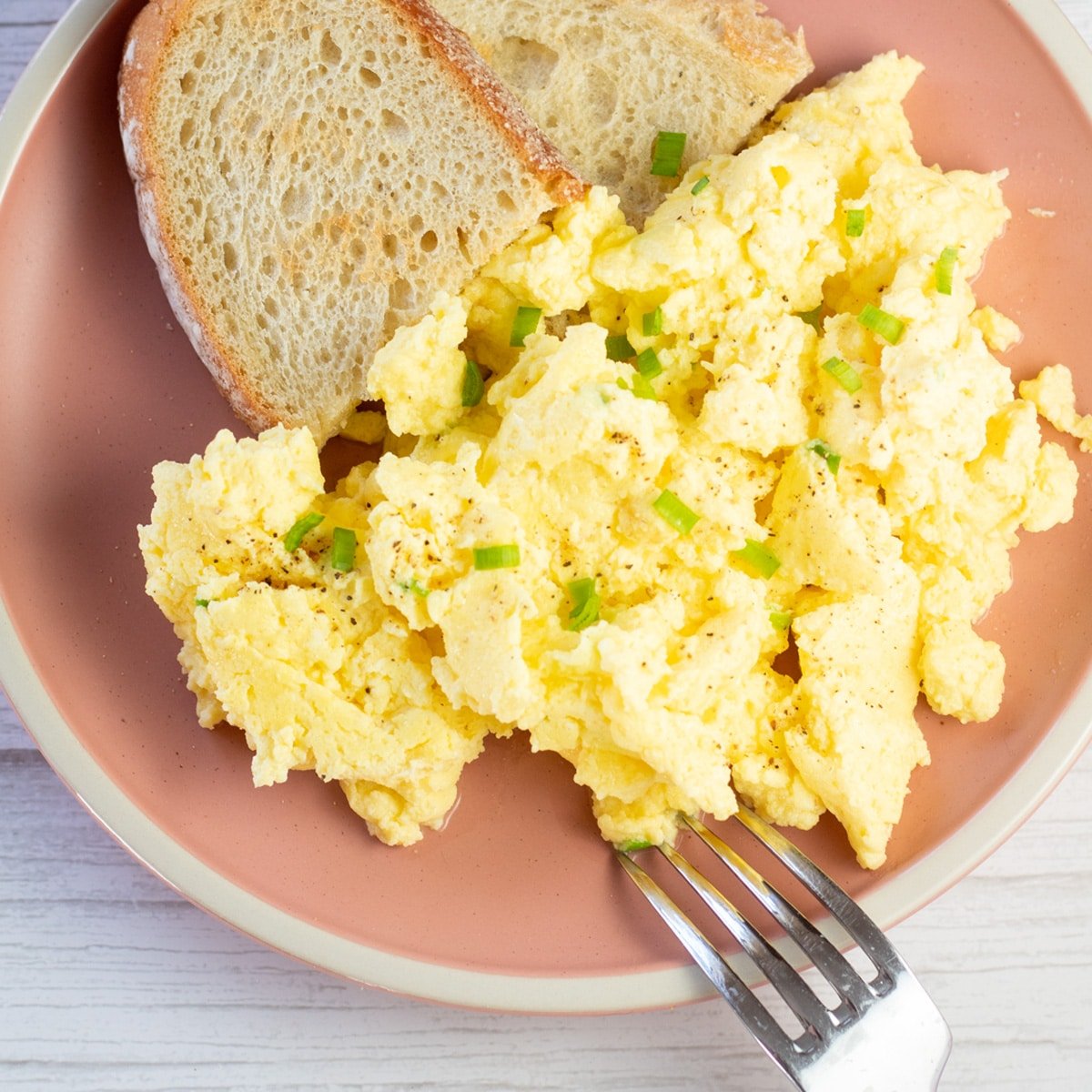 Square image of scrambled eggs on rose colored plate with toast on the side.