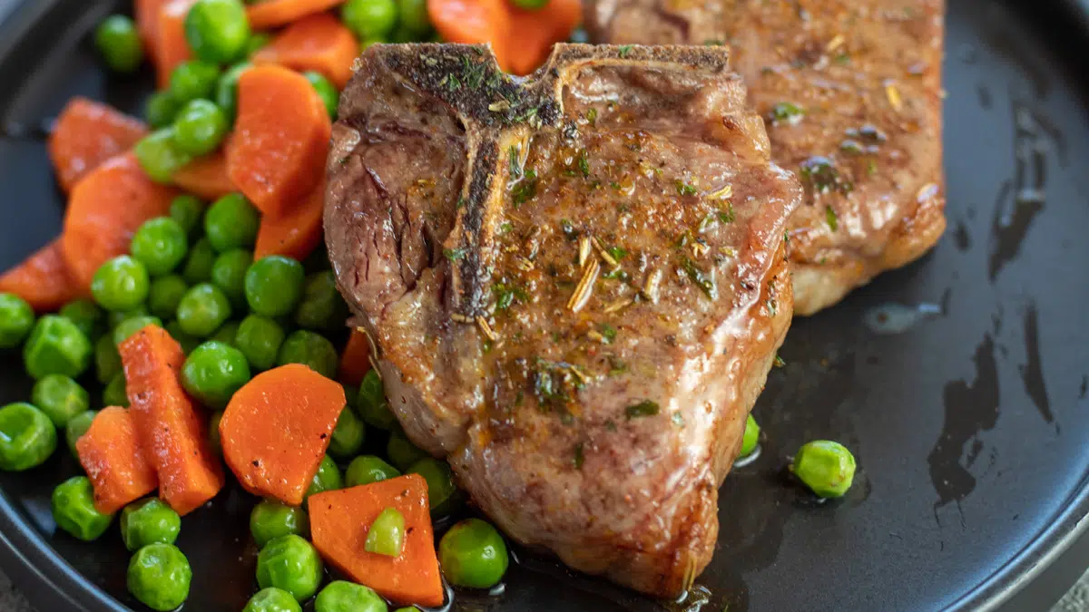 Wide image of air fryer lamb chops on a black plate with peas and carrots on the side.