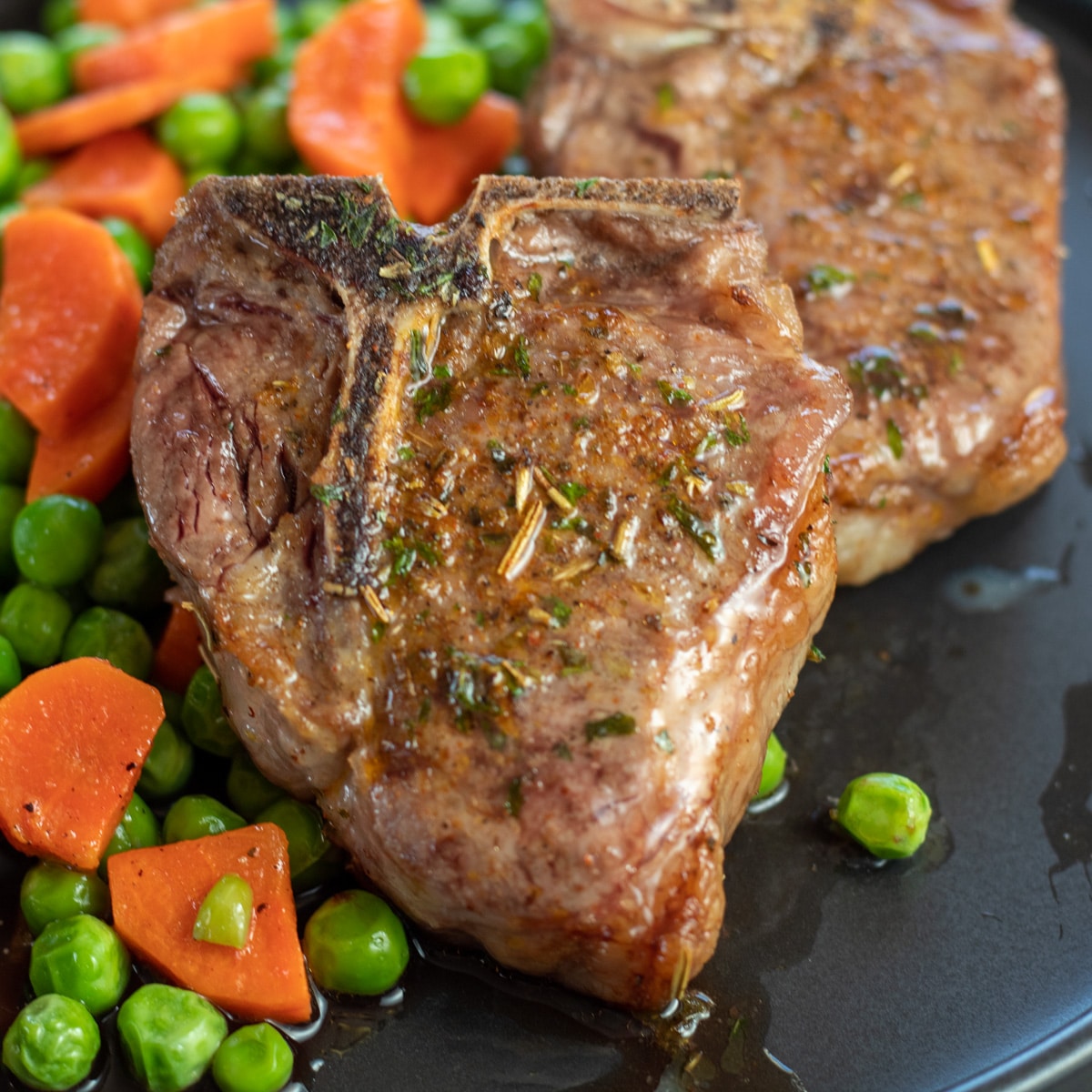 Square image of air fryer lamb chops on a black plate with peas and carrots on the side.