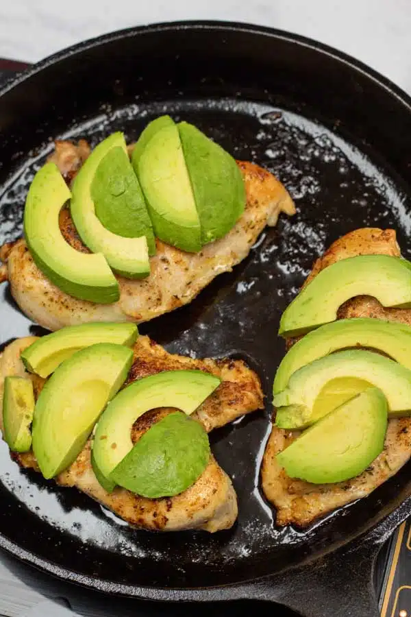 Process photo 5 showing cooked chicken breasts topped with sliced avocado in cast iron pan.