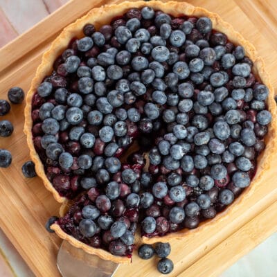 Square image of blueberry tart on a cutting board with a slice being removed.