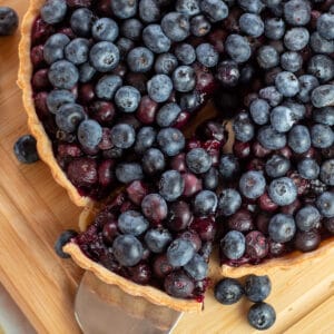 Square close up image of blueberry tart on a cutting board with a slice pulled out.
