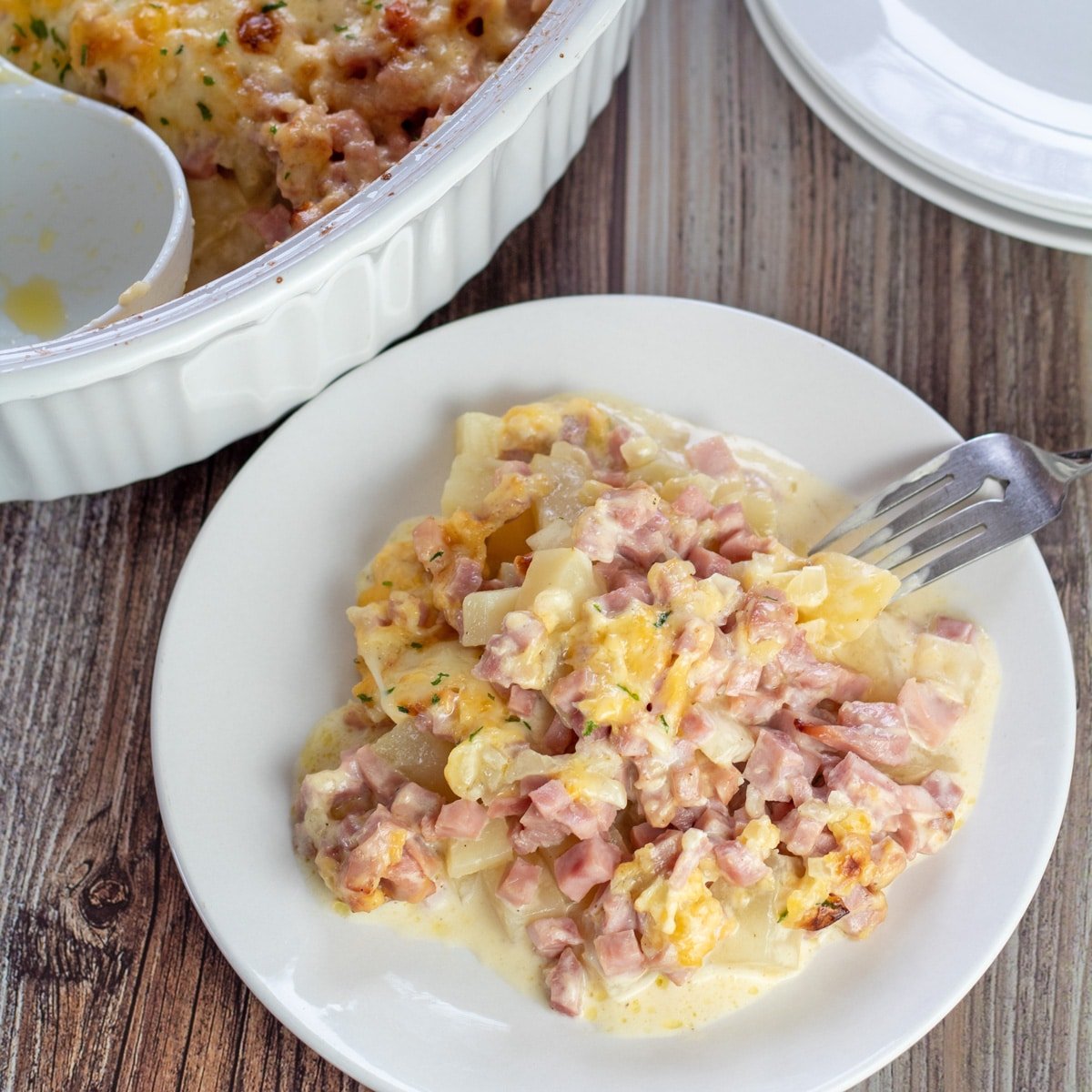 Square overhead image of ham & potato casserole on a white plate, with casserole in background.