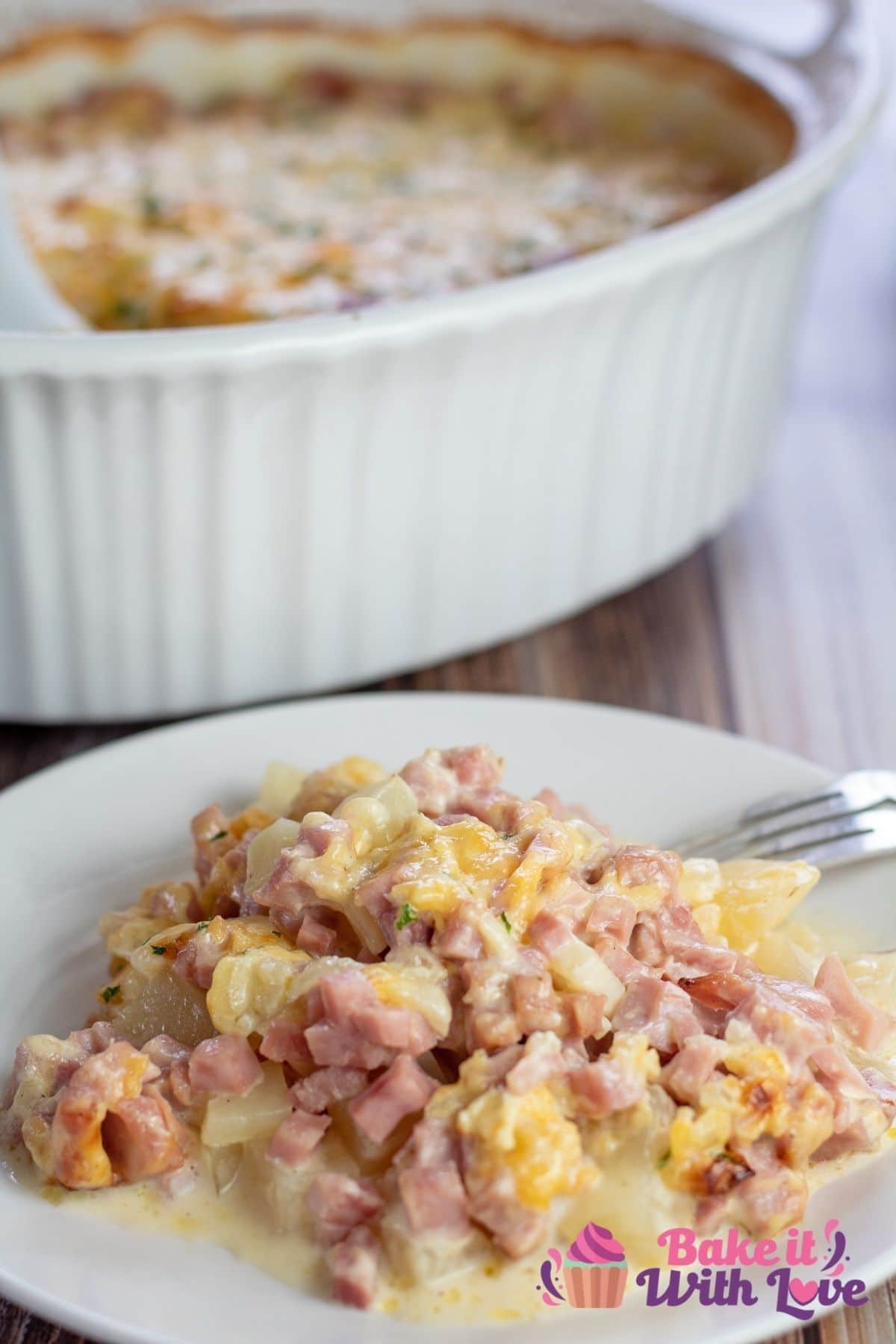 Tall image of ham & potato casserole on a white plate, with casserole in background.