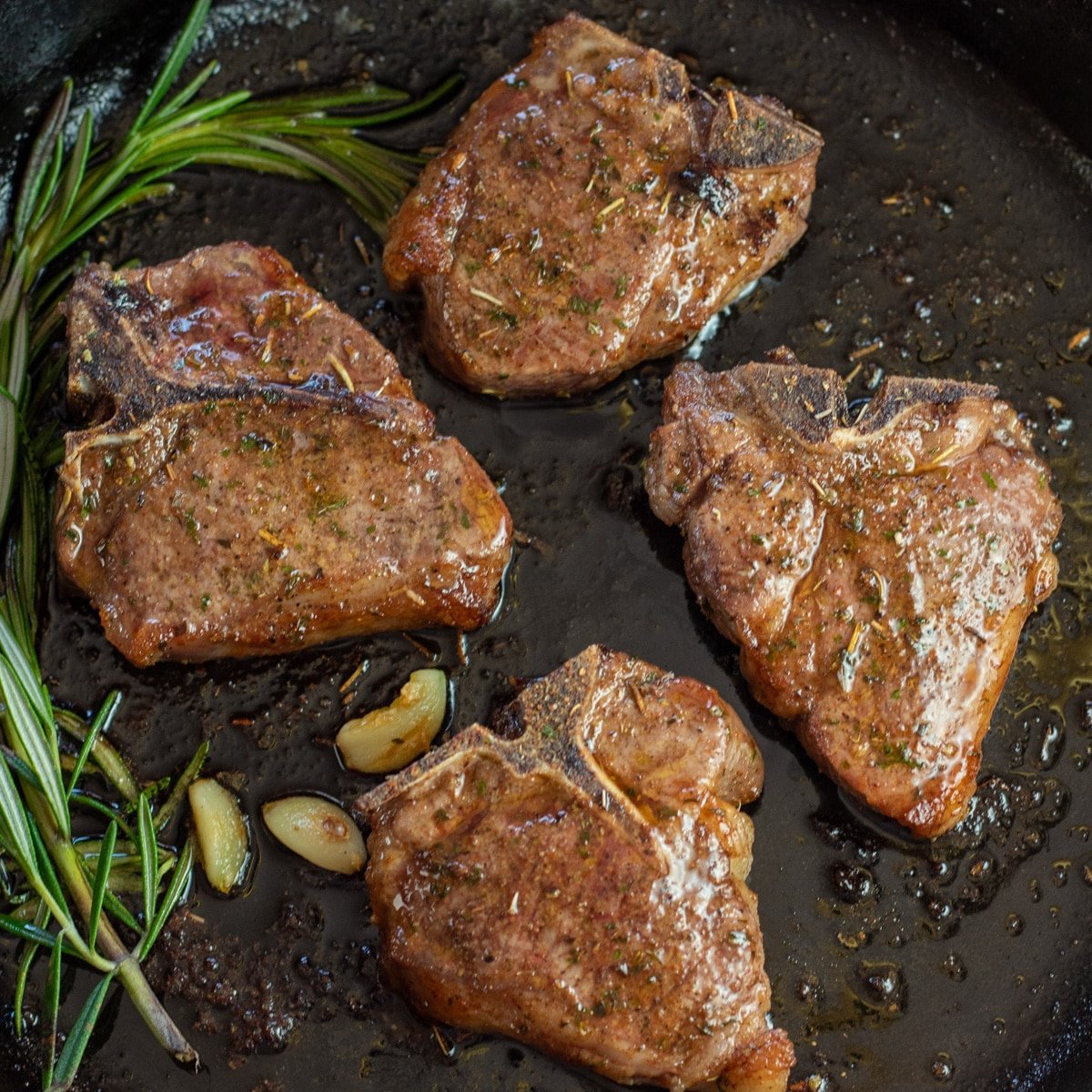 Square image of lamb chops in a cast iron pan with garlic and rosemary.