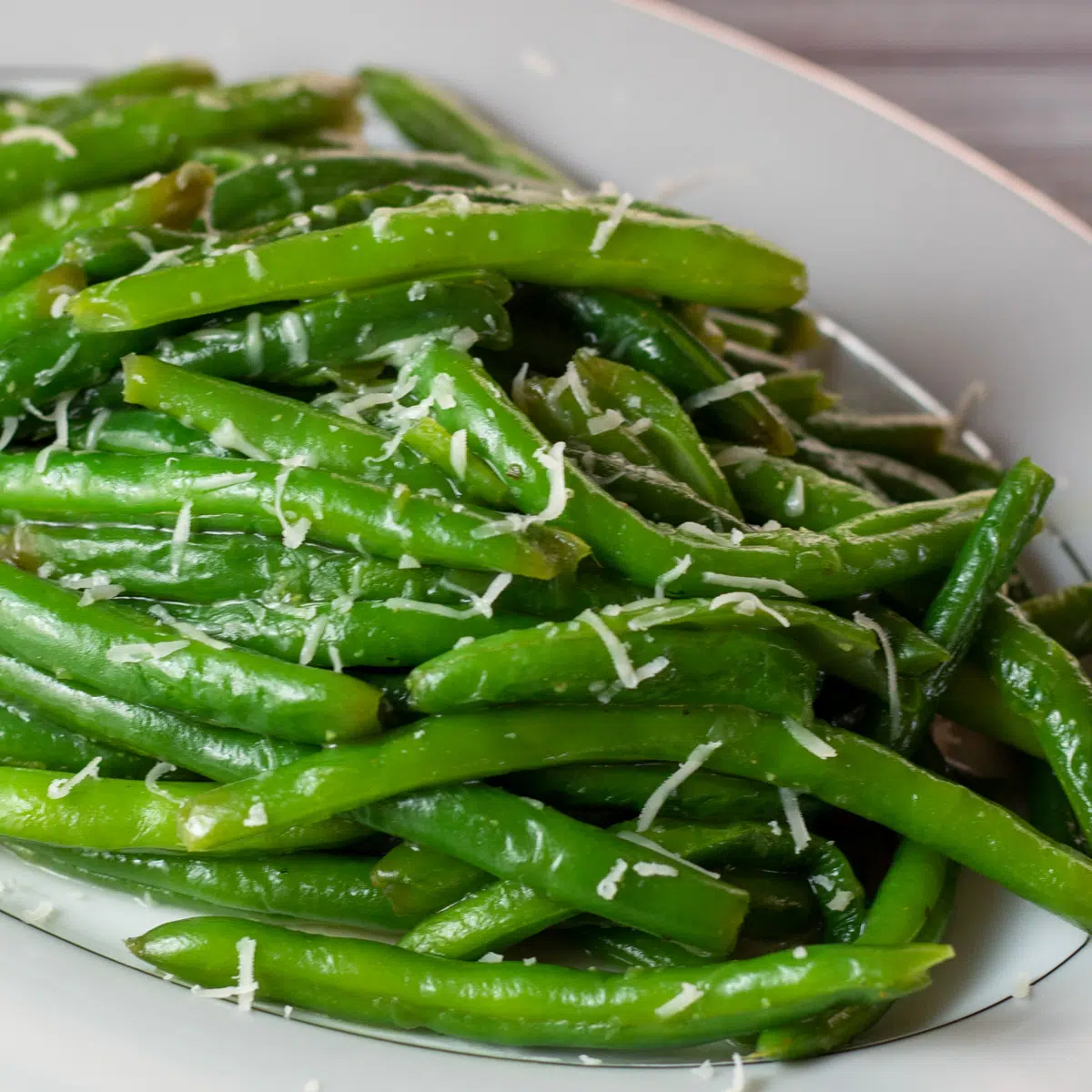Square image of sauteed green beans in a serving dish with Parmesan cheese sprinkled over.
