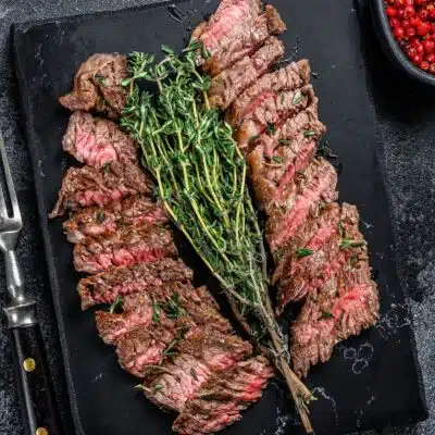 Tender pan seared hanger steak is sliced and served on dark surface with fresh thyme.