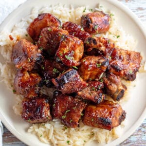 Square image of baked bbq rib tips on a white plate served over rice.