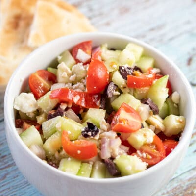 Best Greek salad served in a white bowl with pita bread triangles in background.