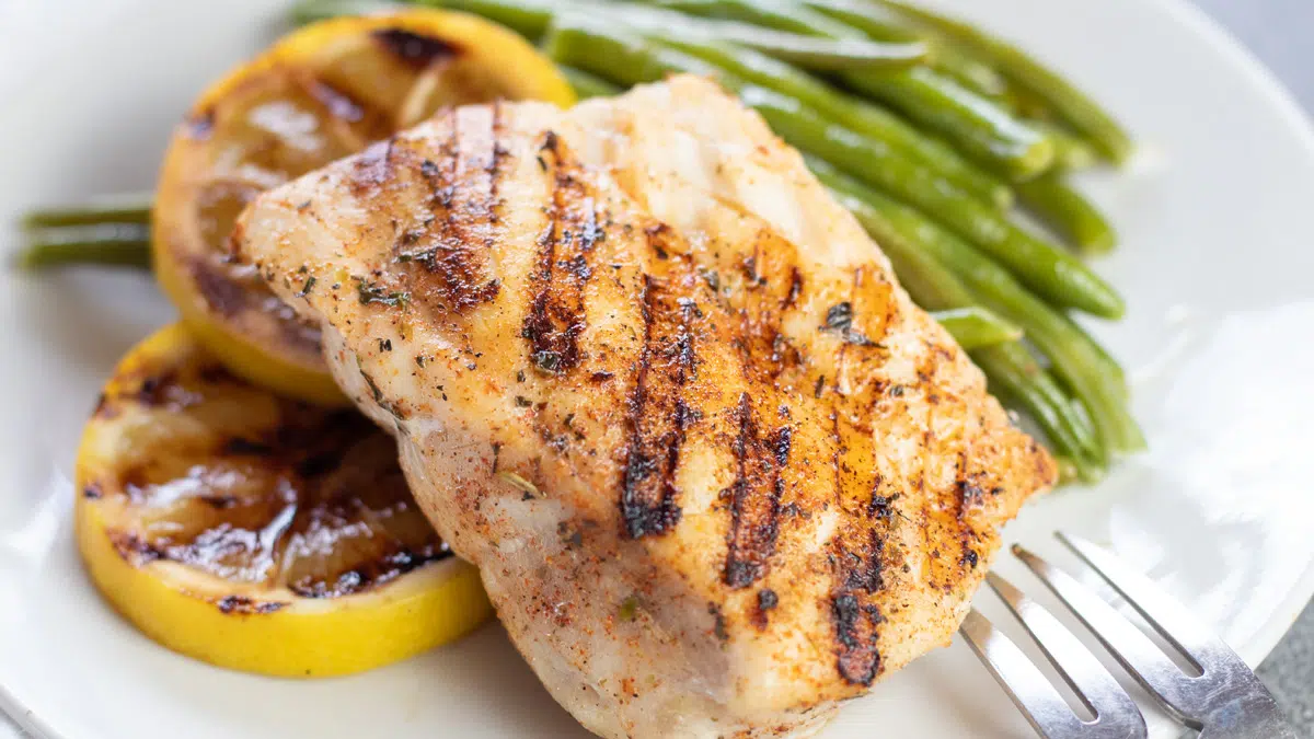 Wide image of grilled grouper fish on a white plate with lemons and green beans.