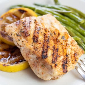 Square image of grilled grouper fish on a white plate with lemons and green beans.