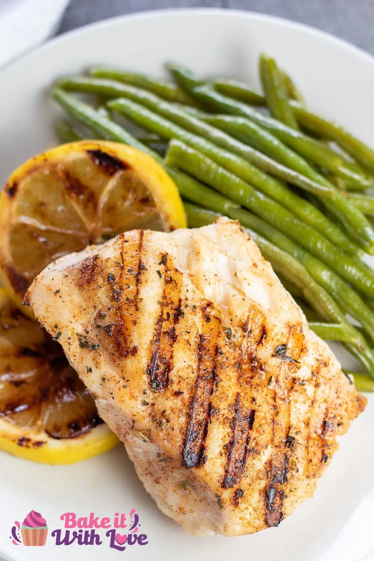 Tall image of grilled grouper fish on a white plate with lemons and green beans.