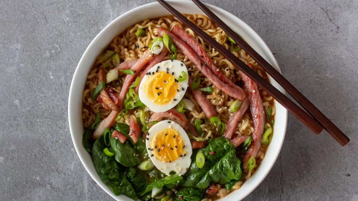 Wide image of ramen noodles with leftover ham and hard boiled eggs.