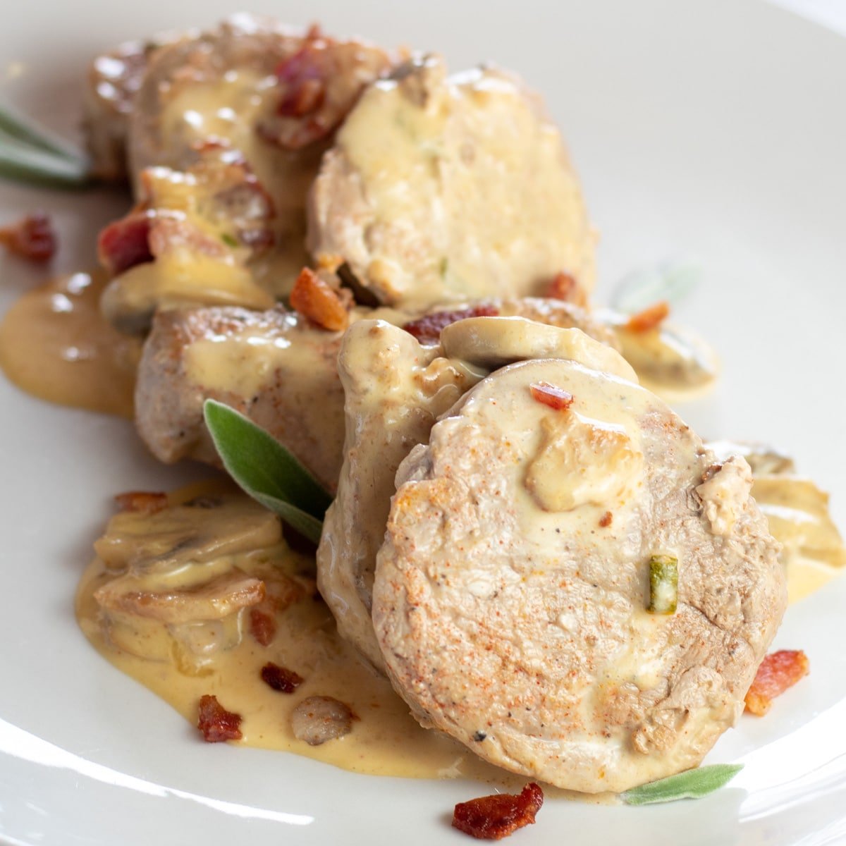 Tenderloin babi dengan saus jamur disajikan di piring putih dan dihiasi dengan daun sage segar.