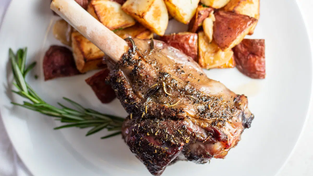 Wide image of roasted lamb shank on a white plate with roasted potatoes.