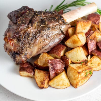 Square image of roasted lamb shank on a white plate with roasted potatoes.