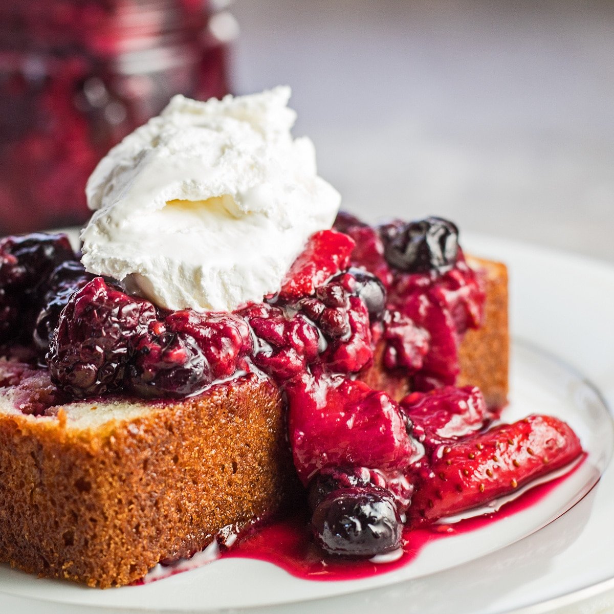 Roasted berries served over raspberry white chocolate loaf cake slice and topped with whipped cream.
