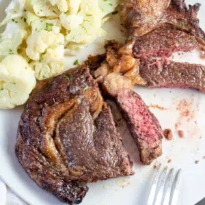 Cut air fryer steak on white plate with cauliflower in background.