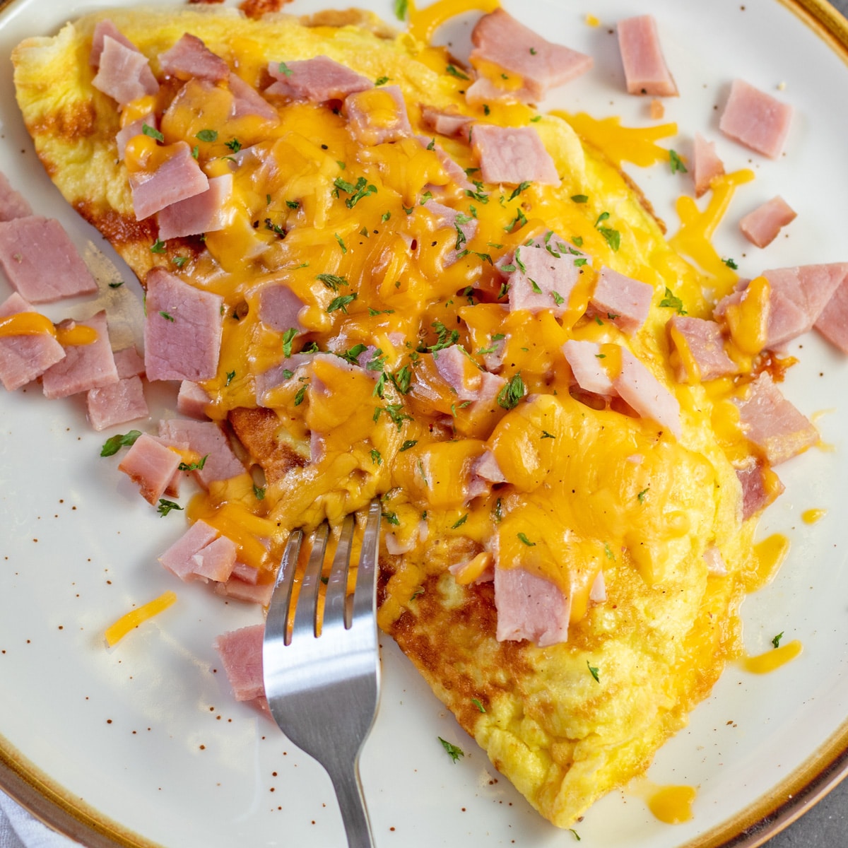 Tasty ham and cheese omelet on tan plate with light brown rim and fork tines near the omelet.