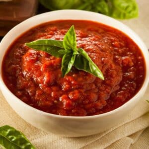 Square image of marinara sauce in a small bowl with a sprig of basil on top.
