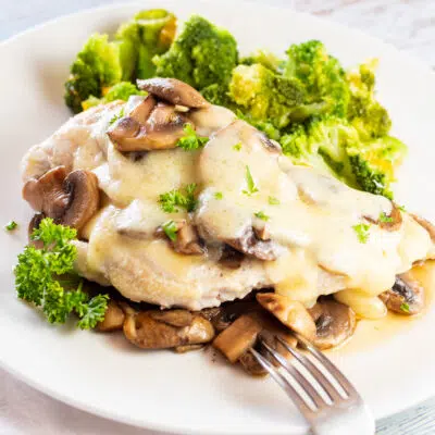 Tender, tasty mushroom swiss chicken is served over more sauteed mushrooms on white plate.